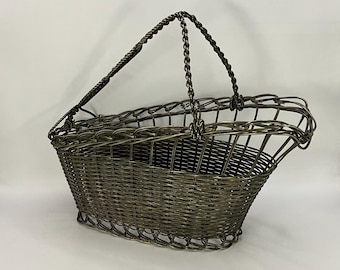 Puede incluir: Una cesta de alambre plateado con un diseño tejido y un asa de alambre trenzado. La cesta tiene forma ovalada y una abertura ancha.