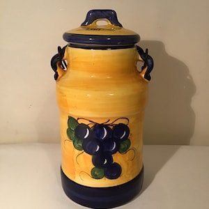 Puede incluir: Un tarro de cerámica amarillo con detalles azules y un diseño de uvas. El tarro tiene dos asas y una tapa.