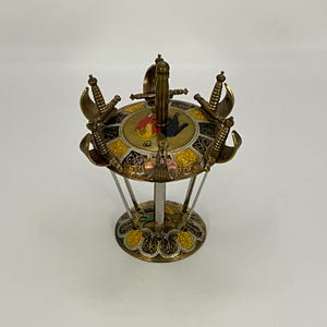 Może przedstawiać: Dekoracyjny uchwyt na wykałaczki z motywem miecza. Uchwyt ma okrągły kształt z centralnym mieczem i mniejszymi mieczami na obwodzie. Schemat kolorów obejmuje złote, czarne, żółte i czerwone akcenty. Jest wspierany przez srebrny stojak.