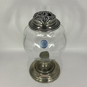 Pode incluir: Um frasco de vidro transparente com tampa e base prateadas. A tampa tem um design floral. O frasco tem um rótulo que diz "Les Etoiles du Loir".