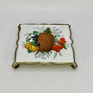 May include: A white ceramic trivet with a gold metal frame. The trivet features a colorful illustration of a pineapple, cherries, and other fruits.