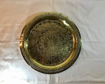 May include: A round, gold-colored metal serving tray with an intricate, embossed floral design. The tray has a raised rim and a smooth, polished surface.