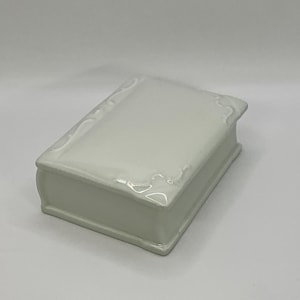 Peut inclure: Une boîte à bijoux blanche en céramique en forme de livre. La boîte a une finition brillante et des détails décoratifs sur le dessus et les bords, ressemblant aux pages d'un livre. La boîte est rectangulaire et repose sur une surface blanche.