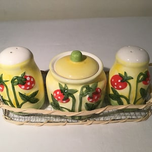May include: A set of three ceramic salt and pepper shakers and a sugar bowl with a wicker tray. The shakers and bowl are decorated with a yellow background and red and white strawberries.