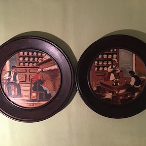 May include: Two round wooden framed plates with painted scenes of people in a shop. The plates are dark brown with a black inner frame. The scenes are painted in a realistic style with muted colors.