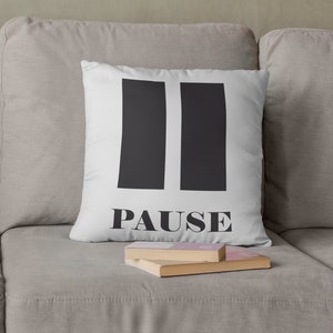 Può includere: Due cuscini bianchi con testo e grafica neri su un divano beige. Un cuscino recita "Nur noch 5 Minuten." L'altro mostra due rettangoli verticali neri con la parola "PAUSE" sotto.