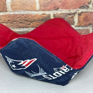 May include: A blue and red fabric bowl with a Patriots logo and a red lining. The bowl is made of fabric and is designed to hold items like snacks or other small items.