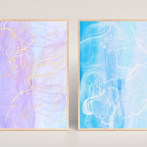 May include: Two framed abstract paintings with a light blue and purple background. The paintings feature white and gold lines.