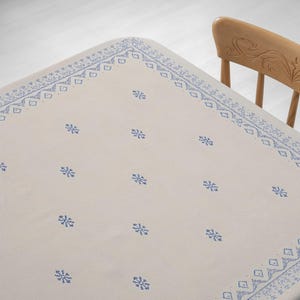 May include: Off-white tablecloth with a blue snowflake pattern and a decorative border. The tablecloth is on a table with a wooden chair in the background. The snowflake pattern is evenly spaced across the surface.