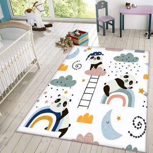Op de afbeelding: Wit tapijt met een pandabeer-, regenboog-, wolken- en sterrenontwerp. Het tapijt is perfect voor een kinderkamer of babykamer.