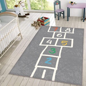 Puede incluir: Una alfombra de hopscotch gris con líneas blancas y números coloridos. La alfombra es perfecta para que los niños jueguen y aprendan sus números.