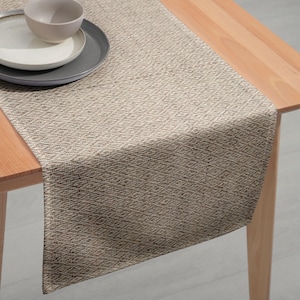 May include: A textured, neutral-toned table runner in beige and brown, draped over a wooden table. The runner's weave creates a subtle pattern. A gray plate, a white plate, and a beige bowl are on the table.