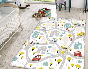 Puede incluir: Una alfombra rectangular con fondo blanco que presenta una red de carreteras gris, casas coloridas en tonos azules y grises, árboles amarillos y varios vehículos, incluyendo un camión de bomberos rojo, un autobús amarillo y un autobús rojo.