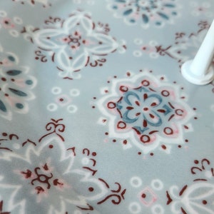 Peut inclure: Gros plan sur un tissu bleu clair avec un motif floral r&eacute;p&eacute;t&eacute; en blanc, rouge fonc&eacute; et bleu clair. Le motif comprend des fleurs stylis&eacute;es et des formes g&eacute;om&eacute;triques. Un objet cylindrique blanc est visible dans le coin sup&eacute;rieur droit.