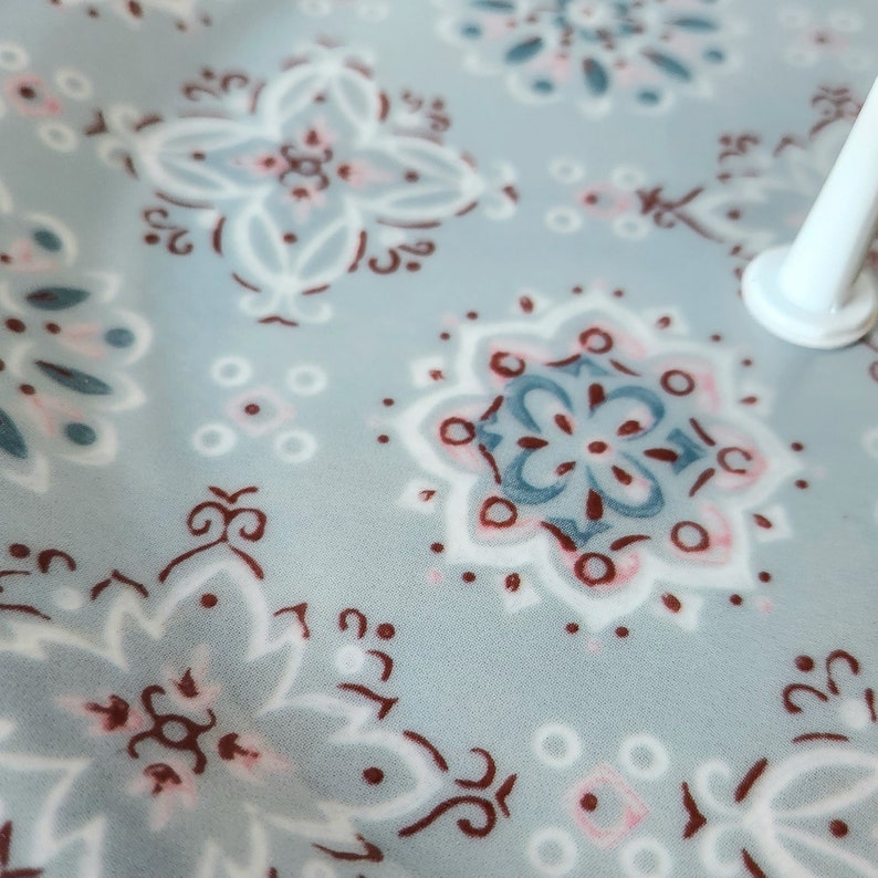 Peut inclure: Gros plan sur un tissu bleu clair avec un motif floral r&eacute;p&eacute;t&eacute; en blanc, rouge fonc&eacute; et bleu clair. Le motif comprend des fleurs stylis&eacute;es et des formes g&eacute;om&eacute;triques. Un objet cylindrique blanc est visible dans le coin sup&eacute;rieur droit.