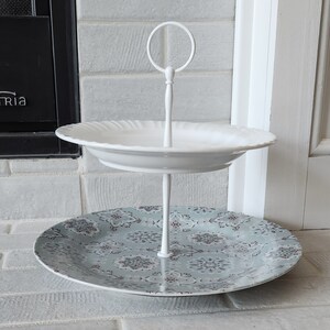 Peut inclure: Pr&eacute;sentoir &agrave; deux niveaux. L'&eacute;tage sup&eacute;rieur est une assiette blanche &agrave; bord festonn&eacute;. L'&eacute;tage inf&eacute;rieur est une assiette &agrave; motifs avec un fond bleu-vert et un motif floral. Le support est blanc et poss&egrave;de une poign&eacute;e en haut.