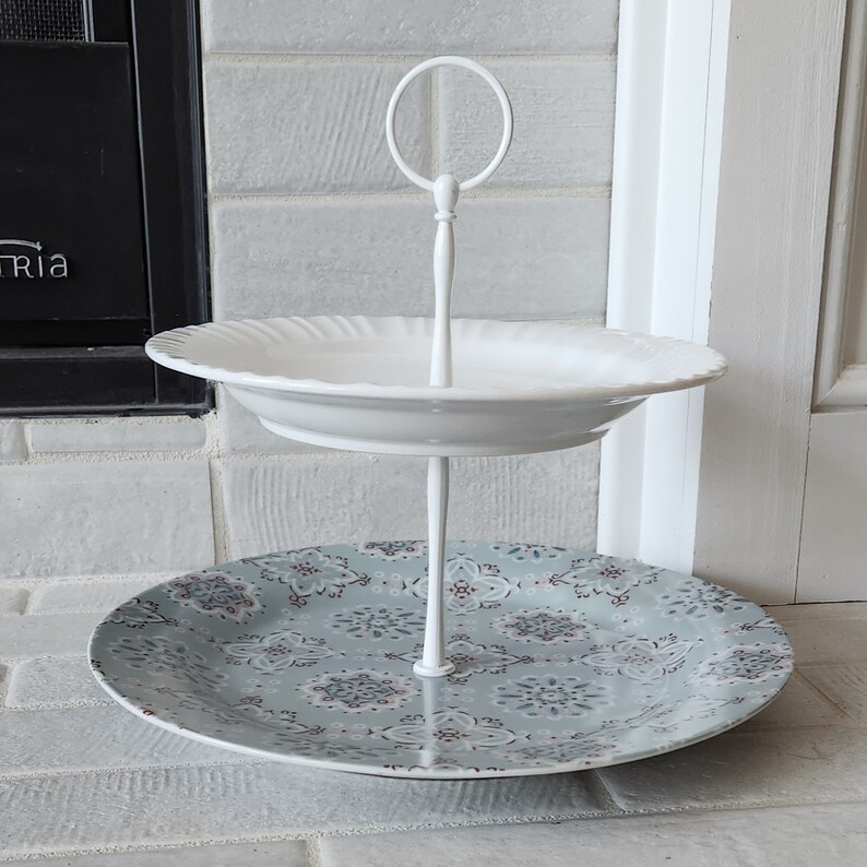 Peut inclure: Pr&eacute;sentoir &agrave; deux niveaux. L'&eacute;tage sup&eacute;rieur est une assiette blanche &agrave; bord festonn&eacute;. L'&eacute;tage inf&eacute;rieur est une assiette &agrave; motifs avec un fond bleu-vert et un motif floral. Le support est blanc et poss&egrave;de une poign&eacute;e en haut.