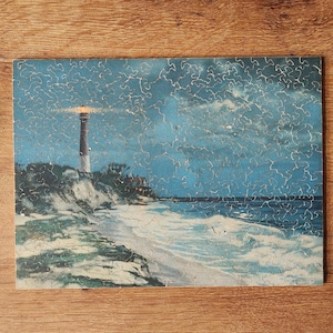 May include: A vintage jigsaw puzzle depicting a lighthouse on a sandy beach with waves crashing on the shore. The lighthouse is lit up at night, and the sky is a dark blue with clouds.