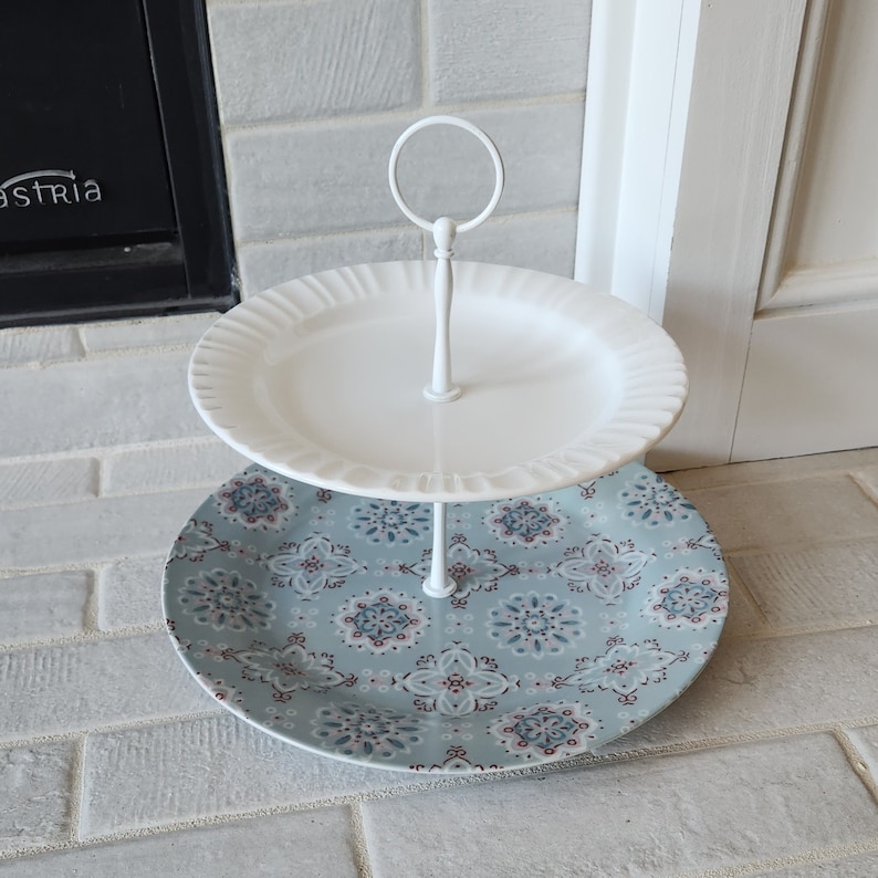Peut inclure: Pr&eacute;sentoir &agrave; deux niveaux avec une structure en m&eacute;tal blanc. Le niveau sup&eacute;rieur est une assiette blanche &agrave; bord festonn&eacute;. Le niveau inf&eacute;rieur est une assiette avec un motif floral bleu et blanc. Id&eacute;al pour les desserts.