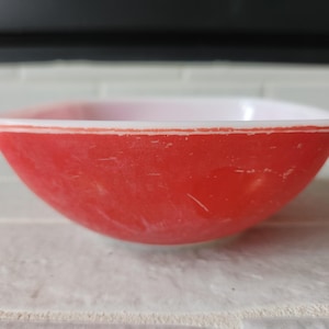 May include: A red and white vintage bowl with a square shape. The bowl has a red exterior with a white interior. A thin orange stripe accents the rim. The bowl is sitting on a white brick surface.