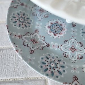 Peut inclure: Une partie d'une assiette en c&eacute;ramique bleu clair avec un motif floral blanc, sarcelle et bordeaux. Le motif de l'assiette pr&eacute;sente des formes de fleurs stylis&eacute;es et des accents g&eacute;om&eacute;triques. Un objet blanc est partiellement visible au-dessus.