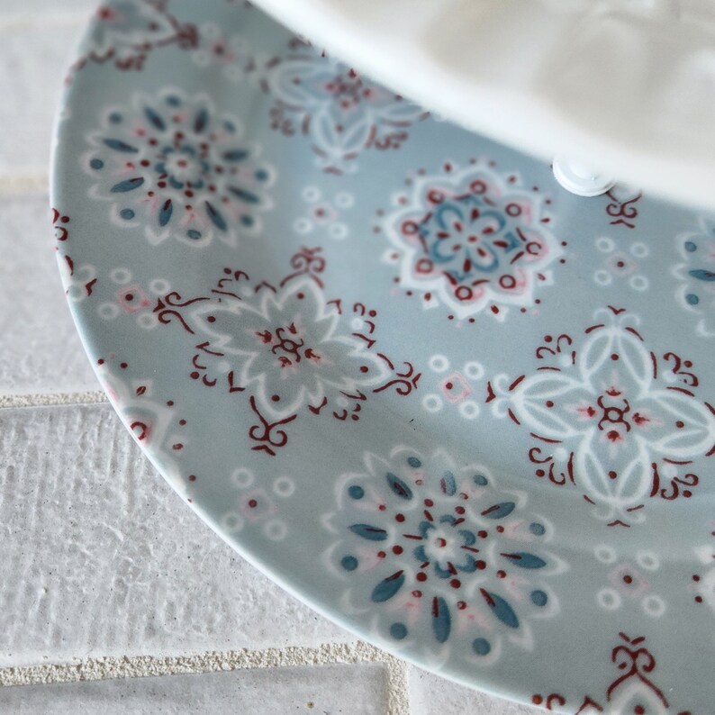 Peut inclure: Une partie d'une assiette en c&eacute;ramique bleu clair avec un motif floral blanc, sarcelle et bordeaux. Le motif de l'assiette pr&eacute;sente des formes de fleurs stylis&eacute;es et des accents g&eacute;om&eacute;triques. Un objet blanc est partiellement visible au-dessus.