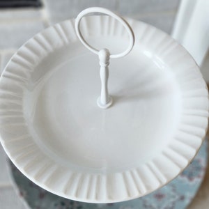 Peut inclure: Un pr&eacute;sentoir &agrave; deux niveaux blanc avec un bord cannel&eacute;. Le niveau sup&eacute;rieur a une poign&eacute;e pour faciliter le transport. Le pr&eacute;sentoir est en c&eacute;ramique ou en porcelaine et est id&eacute;al pour servir des desserts ou des ap&eacute;ritifs. Le plateau mesure environ 25 cm de diam&egrave;tre.