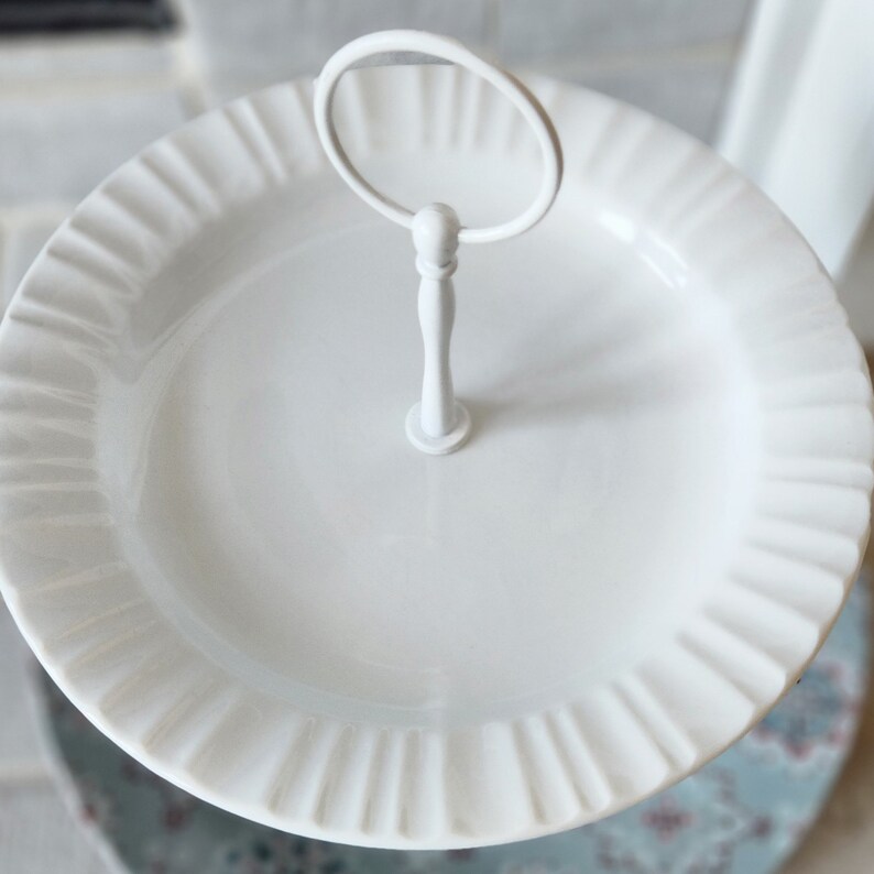Peut inclure: Un pr&eacute;sentoir &agrave; deux niveaux blanc avec un bord cannel&eacute;. Le niveau sup&eacute;rieur a une poign&eacute;e pour faciliter le transport. Le pr&eacute;sentoir est en c&eacute;ramique ou en porcelaine et est id&eacute;al pour servir des desserts ou des ap&eacute;ritifs. Le plateau mesure environ 25 cm de diam&egrave;tre.