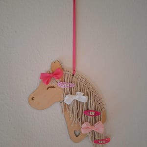 K&ouml;nnte beinhalten: Ein h&ouml;lzerner Einhornkopf mit einem rosa Band und einem Herz-Ausschnitt. Die M&auml;hne des Einhorns besteht aus wei&szlig;em Garn und es sind rosa und wei&szlig;e Haarklammern daran befestigt.