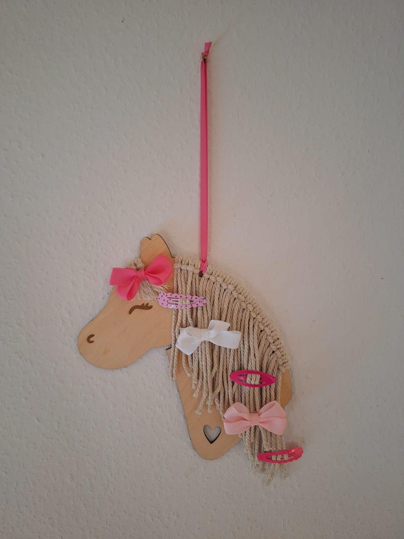 K&ouml;nnte beinhalten: Ein h&ouml;lzerner Einhornkopf mit einem rosa Band und einem Herz-Ausschnitt. Die M&auml;hne des Einhorns besteht aus wei&szlig;em Garn und es sind rosa und wei&szlig;e Haarklammern daran befestigt.