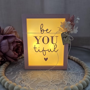 May include: A pink, illuminated box displaying the words "be YOU tiful". The box emits a warm yellow light and is decorated with a small wooden tag with a heart and dried flowers.