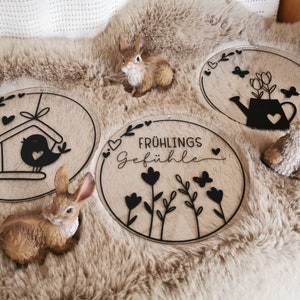 Pu&ograve; includere: Tre cartelli rotondi in legno con disegni in vinile nero. I cartelli mostrano una casetta per uccelli, un annaffiatoio con fiori e un motivo floreale con il testo "Fr&uuml;hlings Gef&uuml;hle".