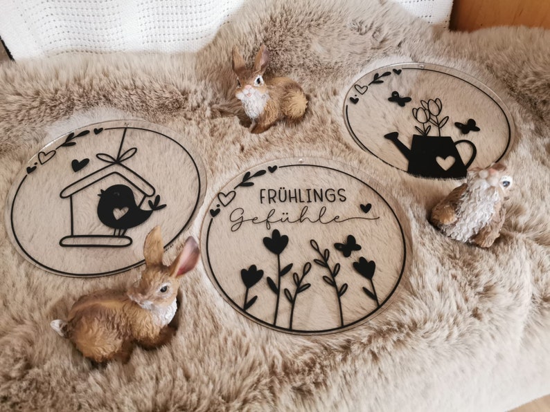 Pu&ograve; includere: Tre cartelli rotondi in legno con disegni in vinile nero. I cartelli mostrano una casetta per uccelli, un annaffiatoio con fiori e un motivo floreale con il testo "Fr&uuml;hlings Gef&uuml;hle".