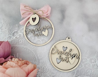 May include: Two wooden ornaments with the words "Erzieherin mit" and "Erzieher mit" written on them. Each ornament has a pink ribbon and three pink hearts.