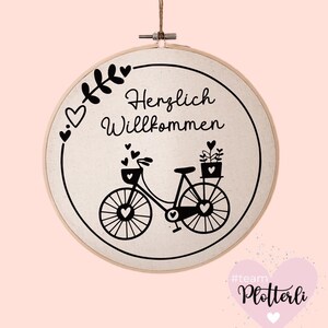 Pode incluir: Bastidor de bordado redondo com tecido creme, apresentando uma ilustra&ccedil;&atilde;o de bicicleta preta com detalhes em forma de cora&ccedil;&atilde;o e a frase alem&atilde; "Herzlich Willkommen". O bastidor &eacute; suspenso por um barbante natural.