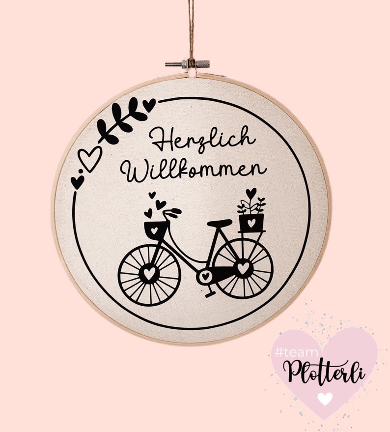 Pode incluir: Bastidor de bordado redondo com tecido creme, apresentando uma ilustra&ccedil;&atilde;o de bicicleta preta com detalhes em forma de cora&ccedil;&atilde;o e a frase alem&atilde; "Herzlich Willkommen". O bastidor &eacute; suspenso por um barbante natural.