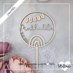 May include: A wooden cake topper featuring the name "Mathilda" and the date "26.10.2024". The design includes a rainbow and the word "TAUFE" on small flags. The topper is circular with a stick for insertion.