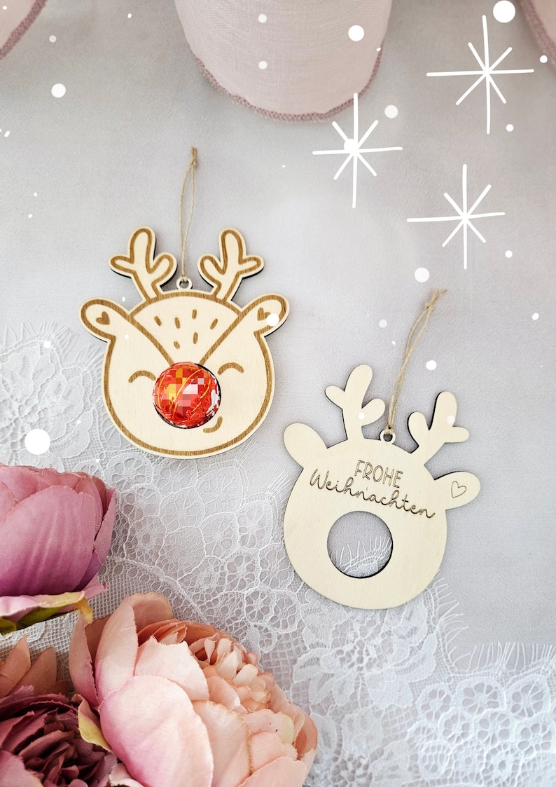 May include: Two wooden Christmas ornaments shaped like reindeer heads. One ornament has a red candy cane in the center of the reindeer's face. The other ornament has the text "Frohe Weihnachten" written on it.