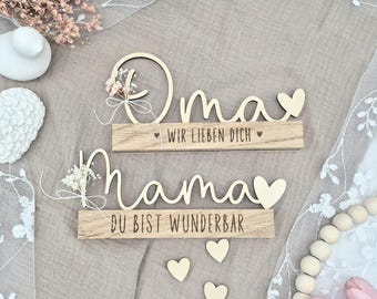 Op de afbeelding: Twee houten borden met de woorden "Oma" en "Mama" in cursief schrift. Het "Oma"-bord heeft de tekst "Wir lieben dich" en het "Mama"-bord heeft de tekst "Du bist wunderbar". Beide borden hebben kleine houten hartjes.