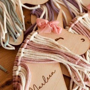 K&ouml;nnte beinhalten: Ein h&ouml;lzerner Einhornkopf mit einer rosa Schleife und einer wei&szlig;en und rosa Garnm&auml;hne. Das Einhorn ist mit dem Namen "Marleen" und einem kleinen Herz-Ausschnitt personalisiert.
