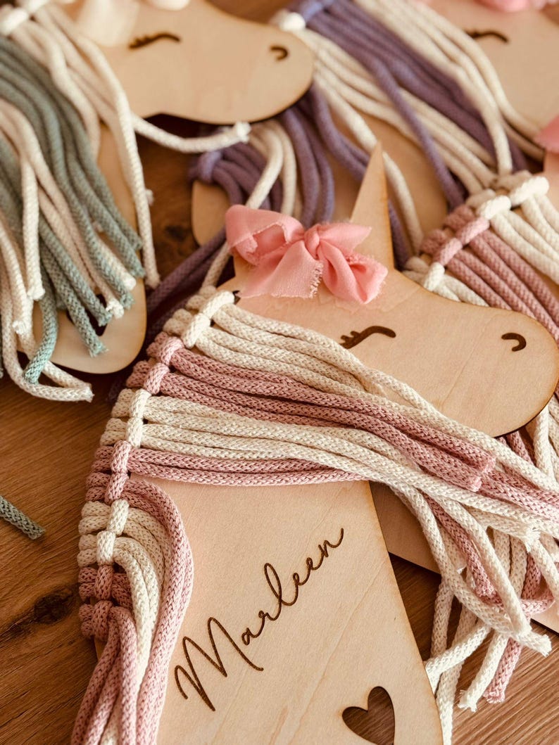 K&ouml;nnte beinhalten: Ein h&ouml;lzerner Einhornkopf mit einer rosa Schleife und einer wei&szlig;en und rosa Garnm&auml;hne. Das Einhorn ist mit dem Namen "Marleen" und einem kleinen Herz-Ausschnitt personalisiert.