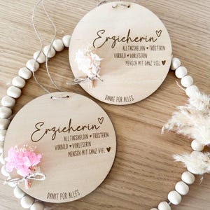 May include: Two round wooden plaques with the word "Erzieherin" and other German text, each adorned with a small bouquet of pink flowers. The plaques are connected by a string of white beads and a natural twine. The text "DANKE FÜR ALLES" is also visible.