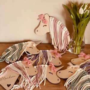K&ouml;nnte beinhalten: Ein Set aus sieben Holz-Einhornkopf-Ornamenten mit buntem Garnhaar. Jedes Einhorn hat eine andere Farbe Garnm&auml;hne und -schwanz. Die Einh&ouml;rner sind in einem Halbkreis auf einer Holzoberfl&auml;che angeordnet. Der Text "Marleen" ist auf einem der Einh&ouml;rner geschrieben.