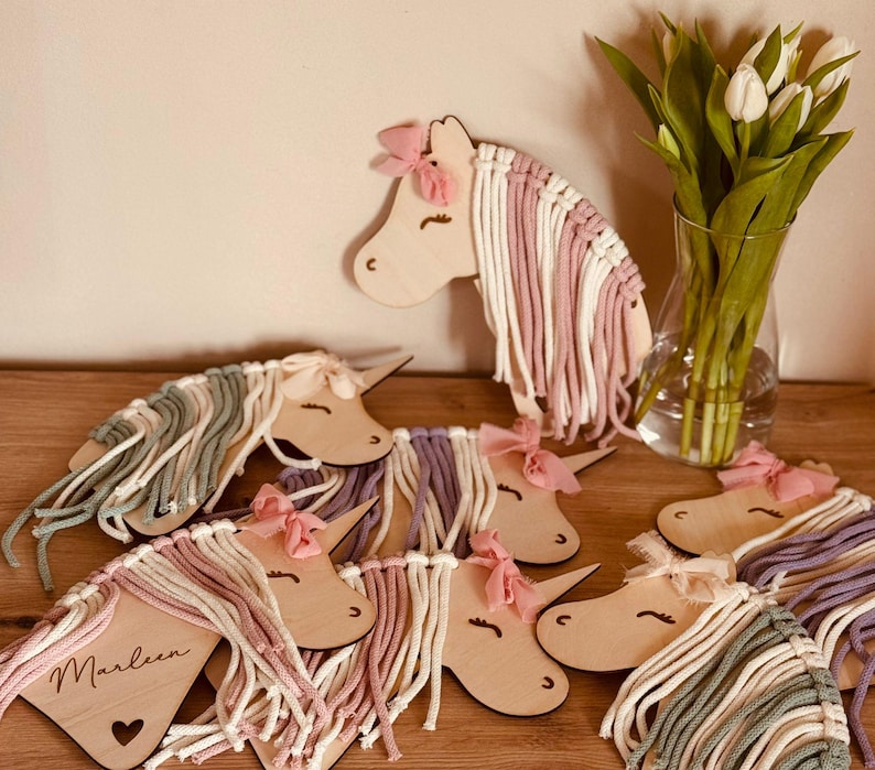 K&ouml;nnte beinhalten: Ein Set aus sieben Holz-Einhornkopf-Ornamenten mit buntem Garnhaar. Jedes Einhorn hat eine andere Farbe Garnm&auml;hne und -schwanz. Die Einh&ouml;rner sind in einem Halbkreis auf einer Holzoberfl&auml;che angeordnet. Der Text "Marleen" ist auf einem der Einh&ouml;rner geschrieben.
