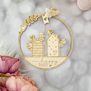 Könnte beinhalten: Holz-Kreis mit einem ausgeschnittenen Design aus zwei Häusern, Grünpflanzen und Herzen. Der Kreis hat eine Schnur daran befestigt und der Text "happy place" steht unten.