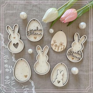 May include: Seven wooden Easter decorations, including bunnies, eggs, and a floral design. The decorations are all white with a natural wood finish and have a cutout design. The eggs have the text "Frohe Ostern" written on them.