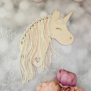 K&ouml;nnte beinhalten: Ein h&ouml;lzerner Einhornkopf mit einer pinkfarbenen und wei&szlig;en Garnm&auml;hne und einem kleinen Herz-Ausschnitt. Das Einhorn befindet sich auf einem wei&szlig;en Spitzenhintergrund mit rosa Blumen.