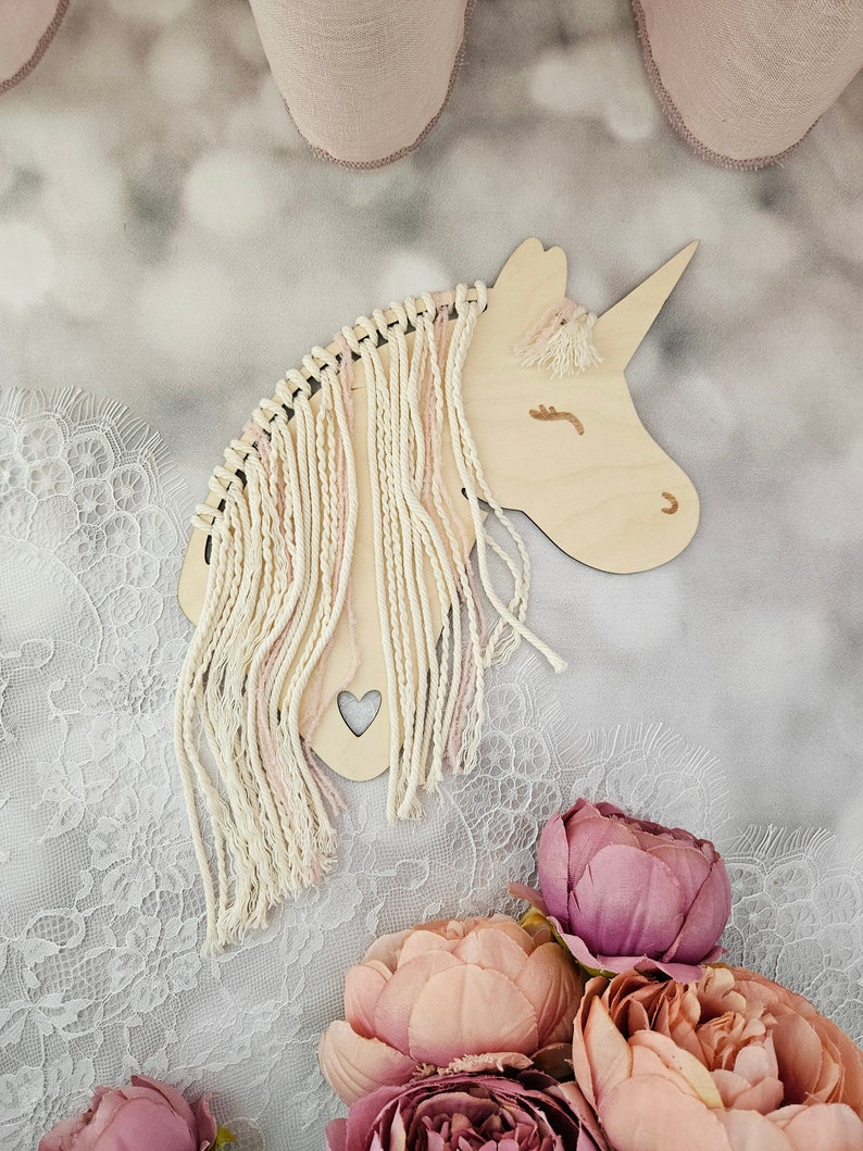 K&ouml;nnte beinhalten: Ein h&ouml;lzerner Einhornkopf mit einer pinkfarbenen und wei&szlig;en Garnm&auml;hne und einem kleinen Herz-Ausschnitt. Das Einhorn befindet sich auf einem wei&szlig;en Spitzenhintergrund mit rosa Blumen.