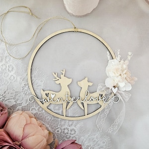 May include: A wooden hoop with a pair of deer silhouettes and the text "winterlove" in cursive. The hoop is decorated with a white lace ribbon and a small bouquet of white flowers.