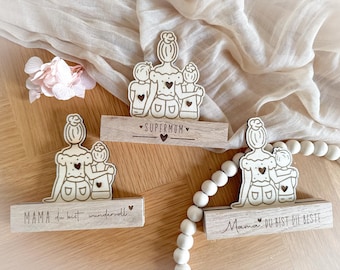 May include: Three wooden cutout figures of a mother and two children, each holding a heart. The figures are standing on a wooden base with the text "SUPERMOM" on the middle base, "MAMA du bist wundervoll" on the left base, and "Mama DU BIST DIE BESTE" on the right base.